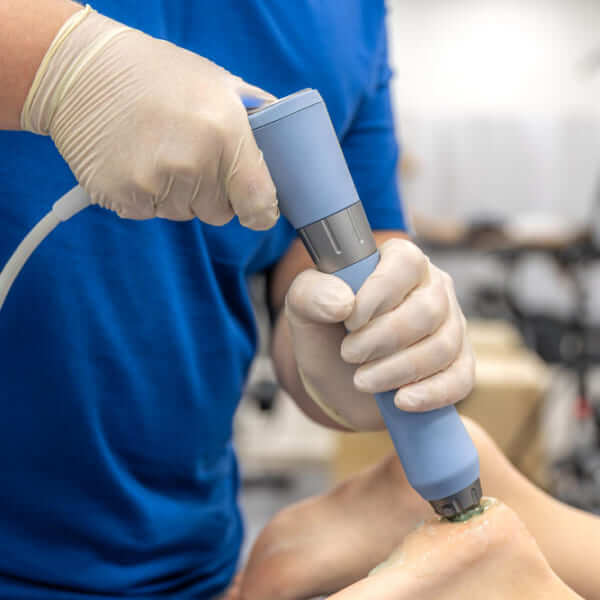 A medical professional wearing tan gloves using a handheld shockwave therapy device on a patient's heel to treat chronic foot pain.