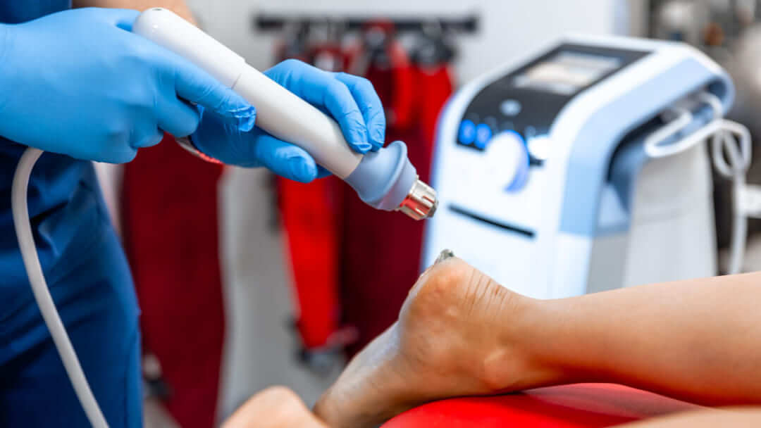 A medical professional wearing tan gloves using a handheld shockwave therapy device on a patient's heel to treat chronic foot pain.